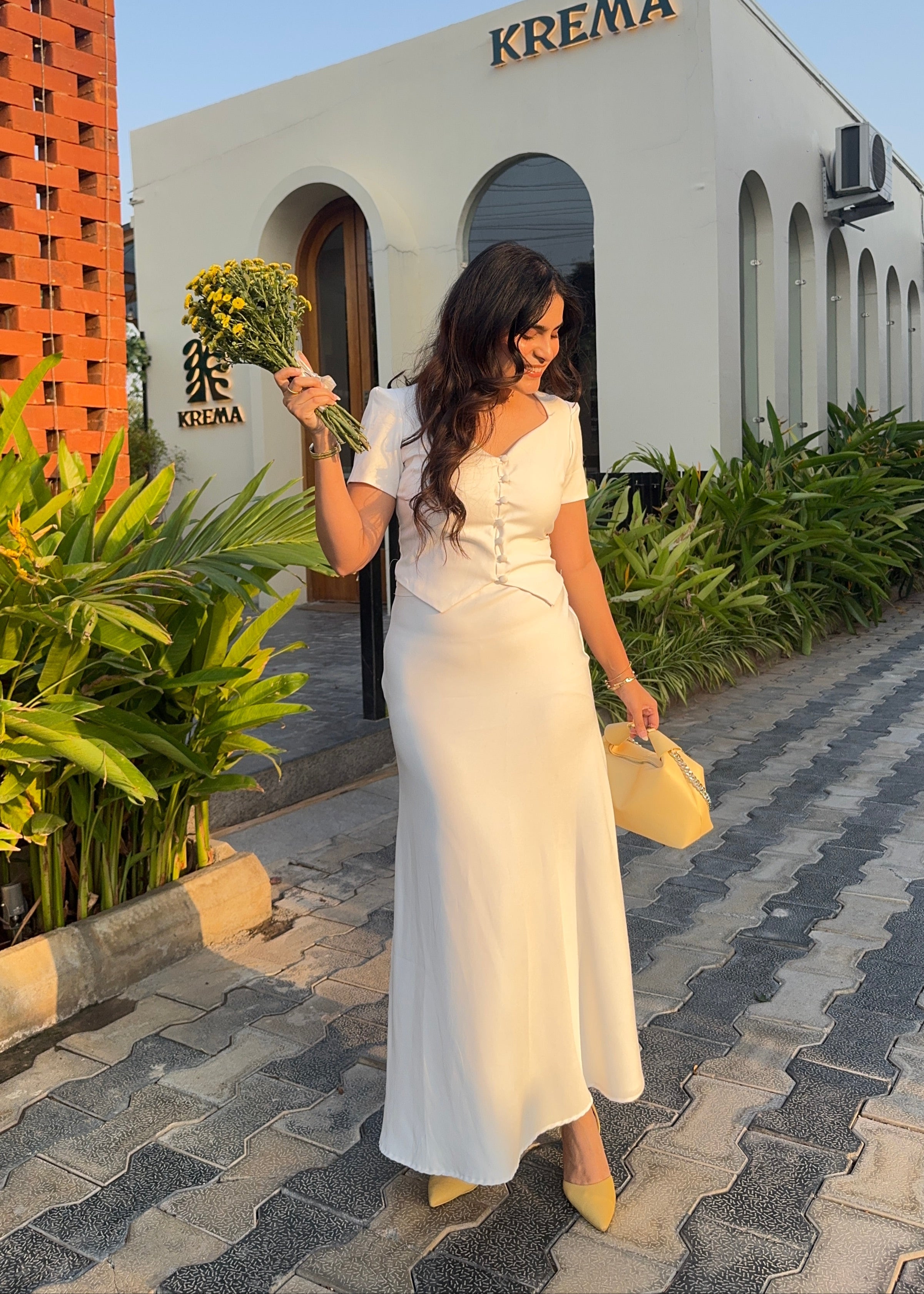 Woman in a white dress holding flowers in front of a building with 'Kremia' branding.