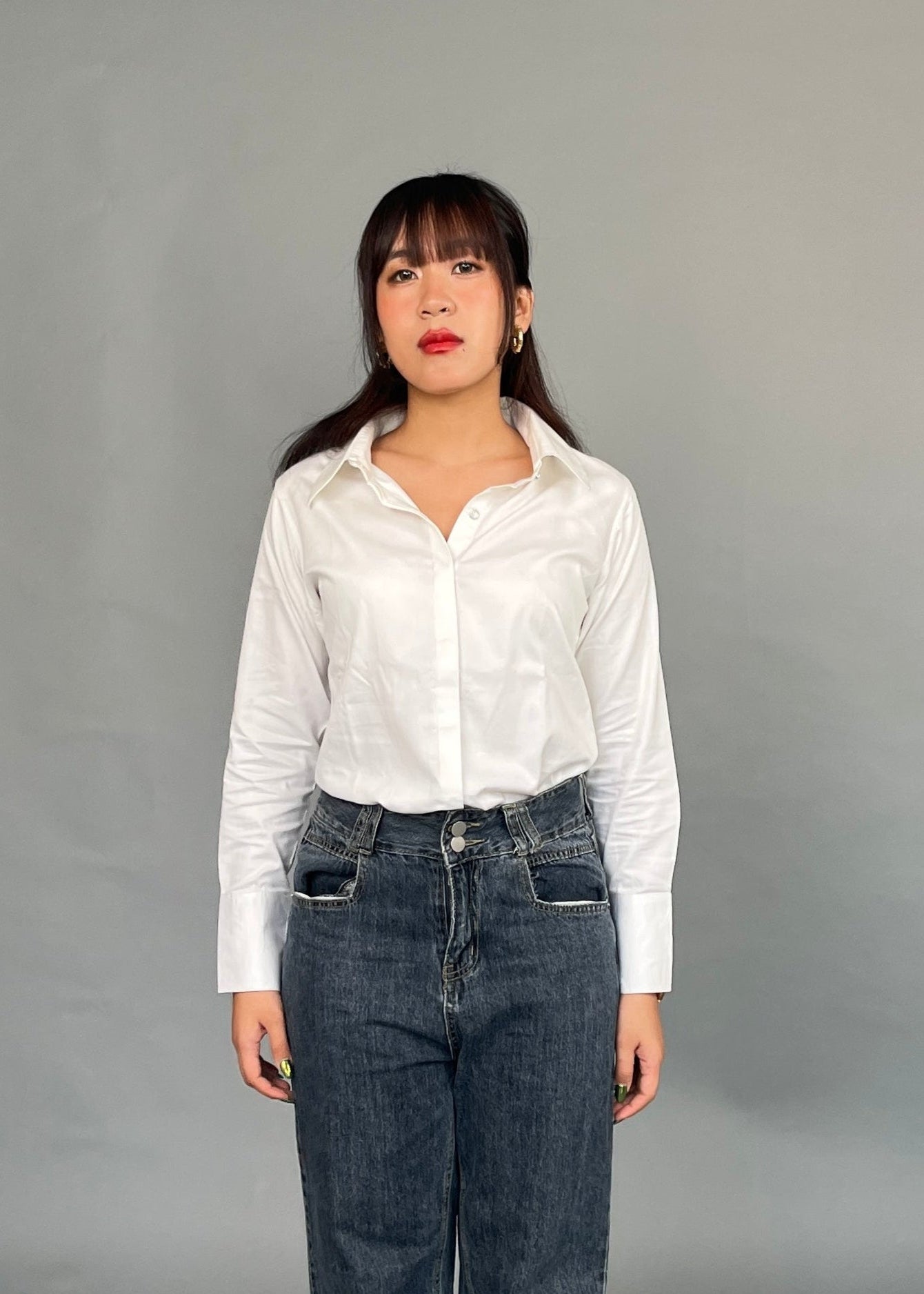A woman standing in a studio setting, wearing a classic white shirt with long sleeves and a collar, paired with blue jeans. She has a neutral expression and is posing with her hands at her sides.