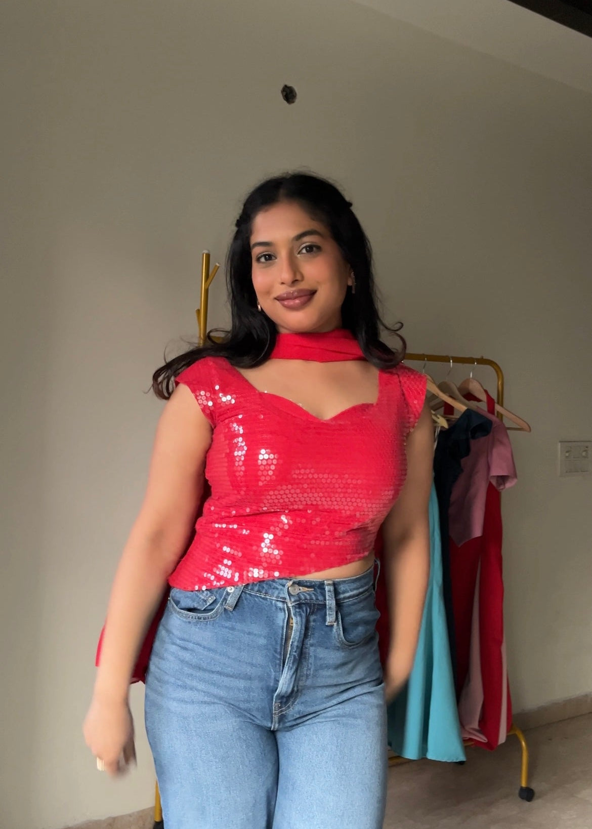 Woman wearing a red top and blue jeans standing in a room with clothes in the background.