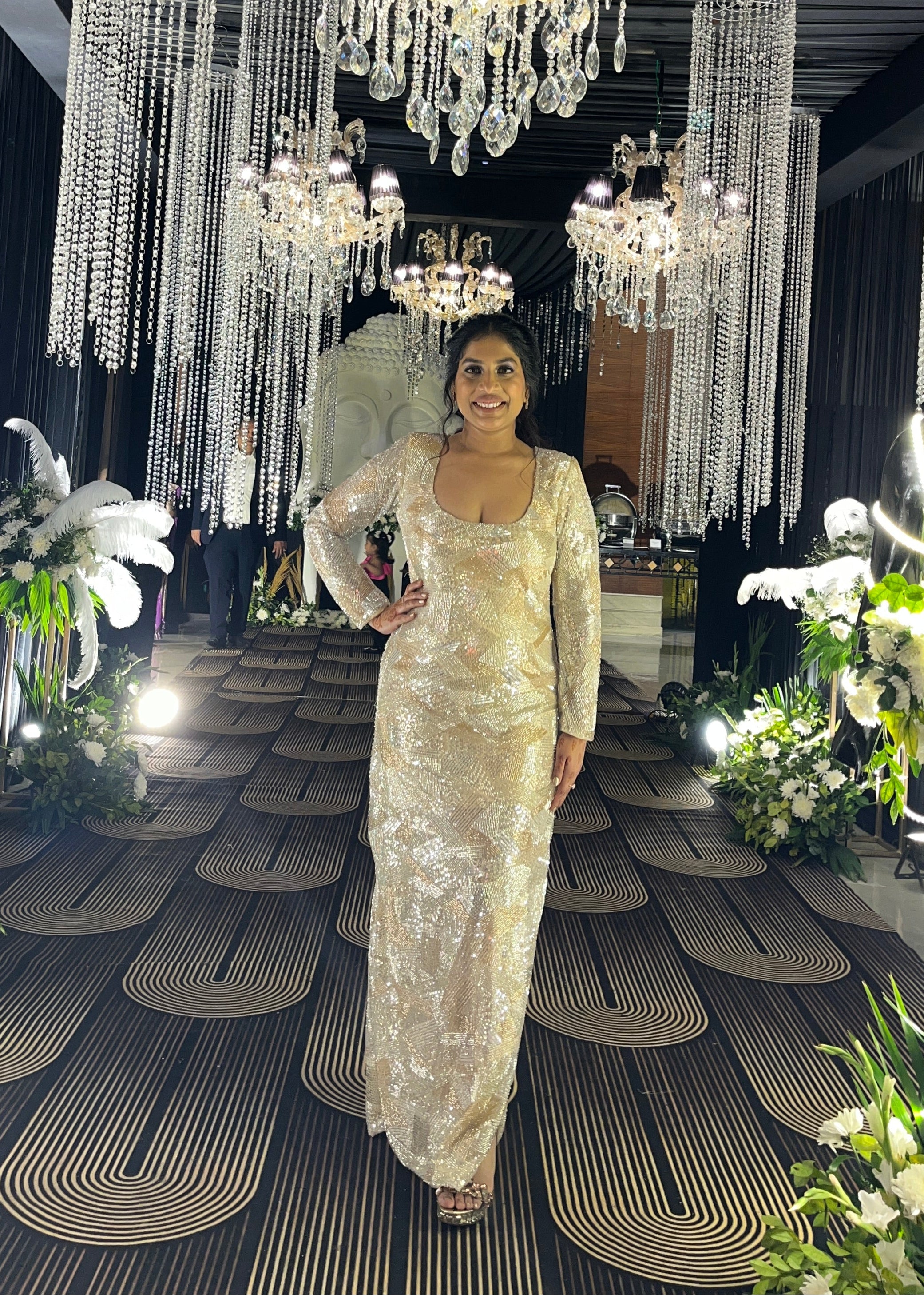 Person in a sparkling dress standing in front of a large chandelier with floral decorations.