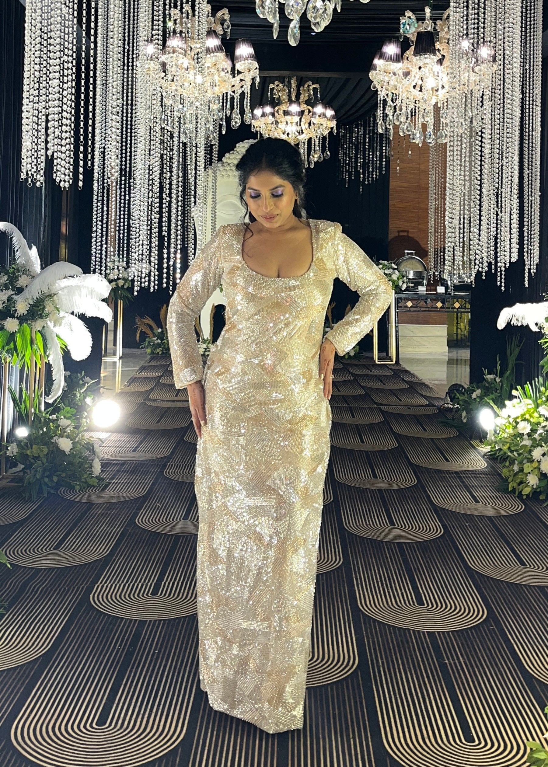 Woman in a sparkling dress standing in an elegant indoor setting with chandeliers and floral arrangements.