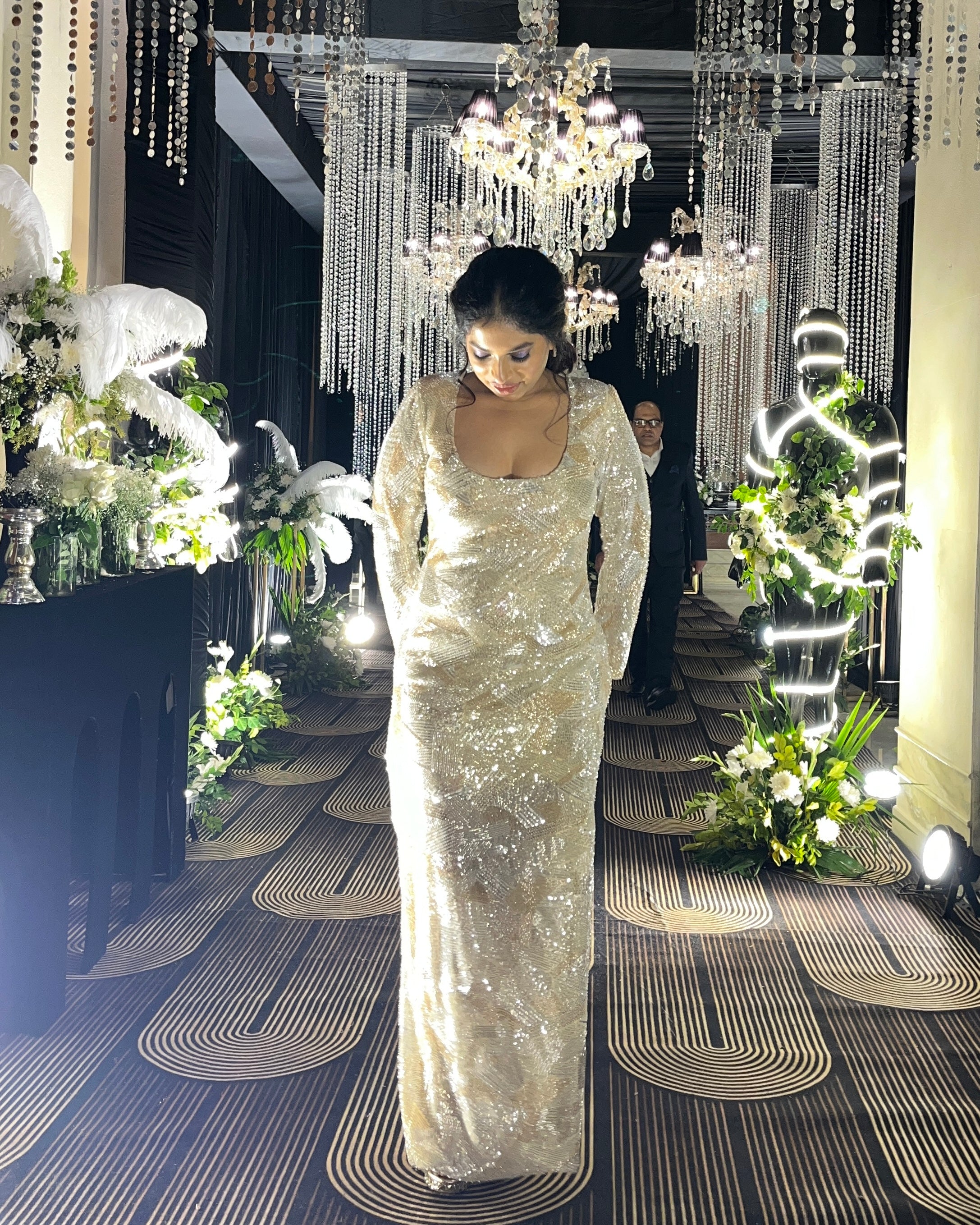 Woman in a sparkling silver gown standing in an opulent room with chandeliers and floral arrangements.