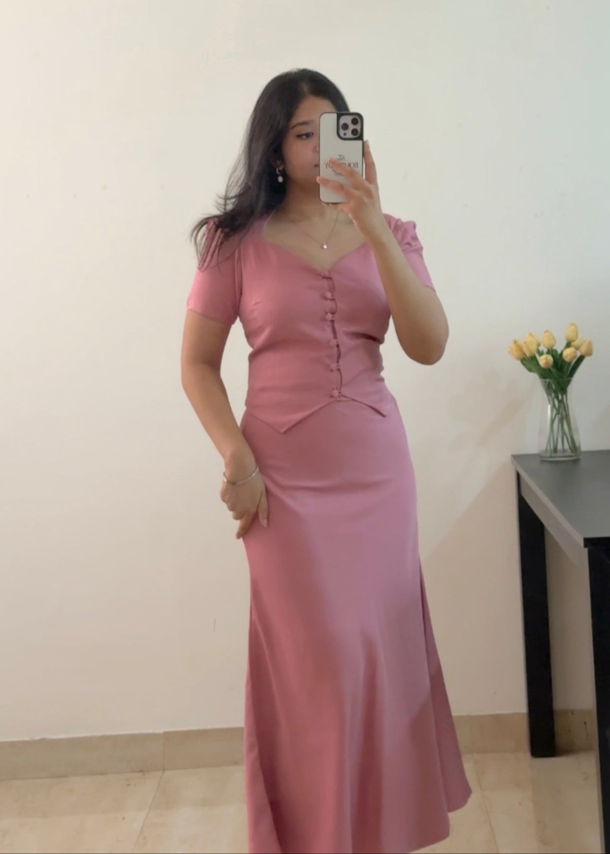 Woman wearing a pink dress taking a mirror selfie in a room with a table and flowers.