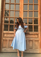 Woman in a light blue dress standing in front of a large wooden door with glass panels.
