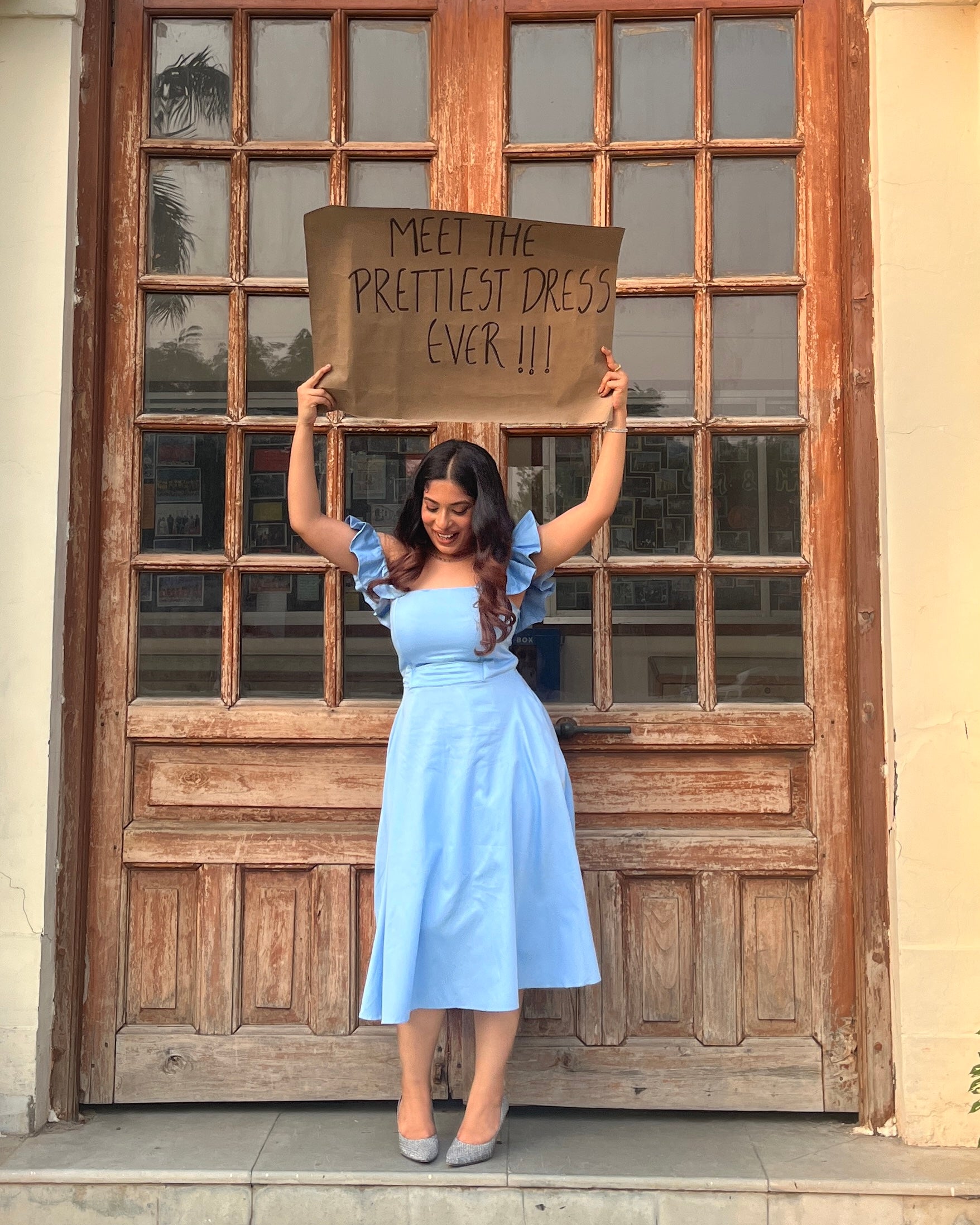 Woman in a light blue dress holding up a cardboard sign in front of a wooden door.