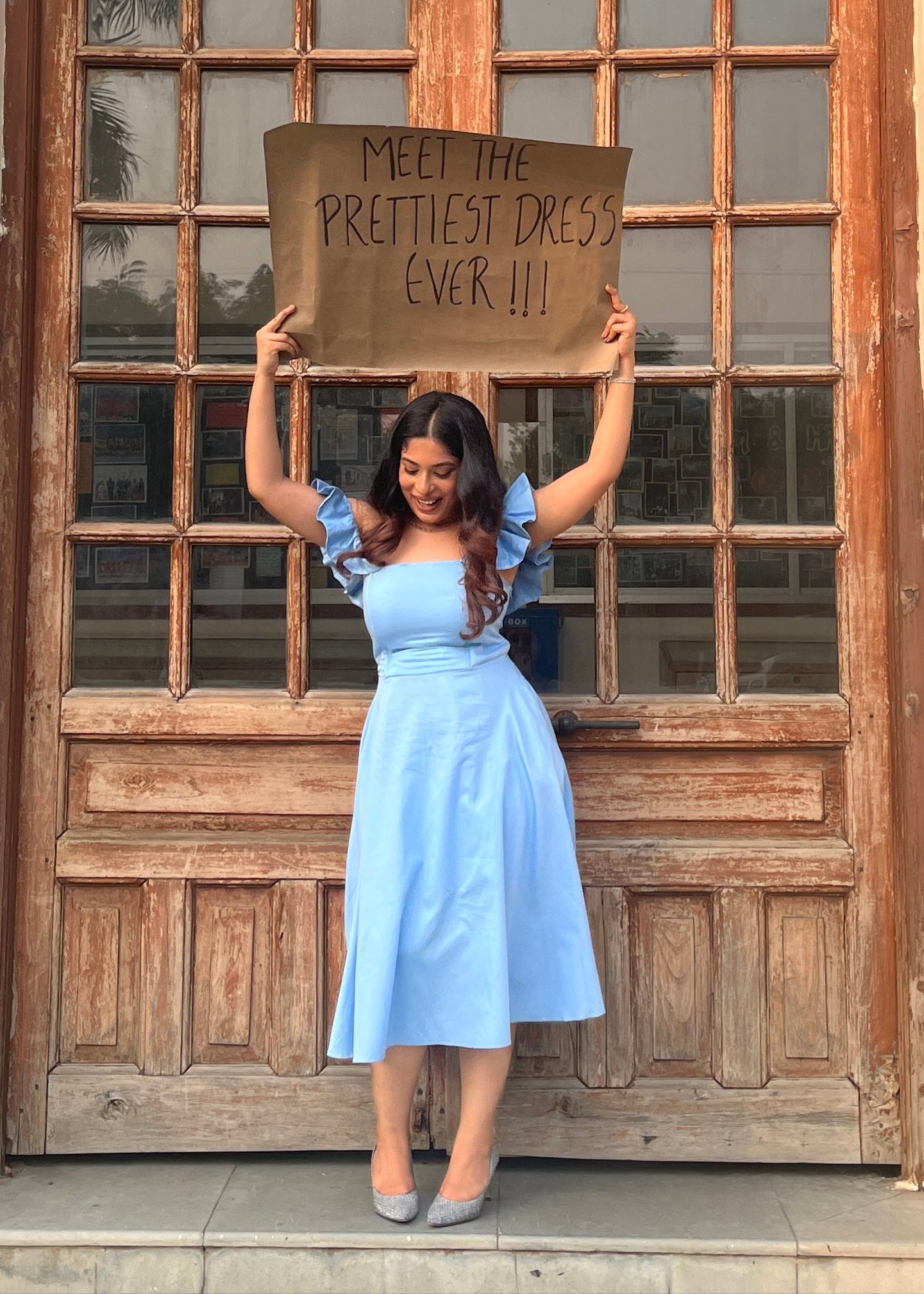 Woman in a light blue dress holding up a cardboard sign in front of a wooden door.