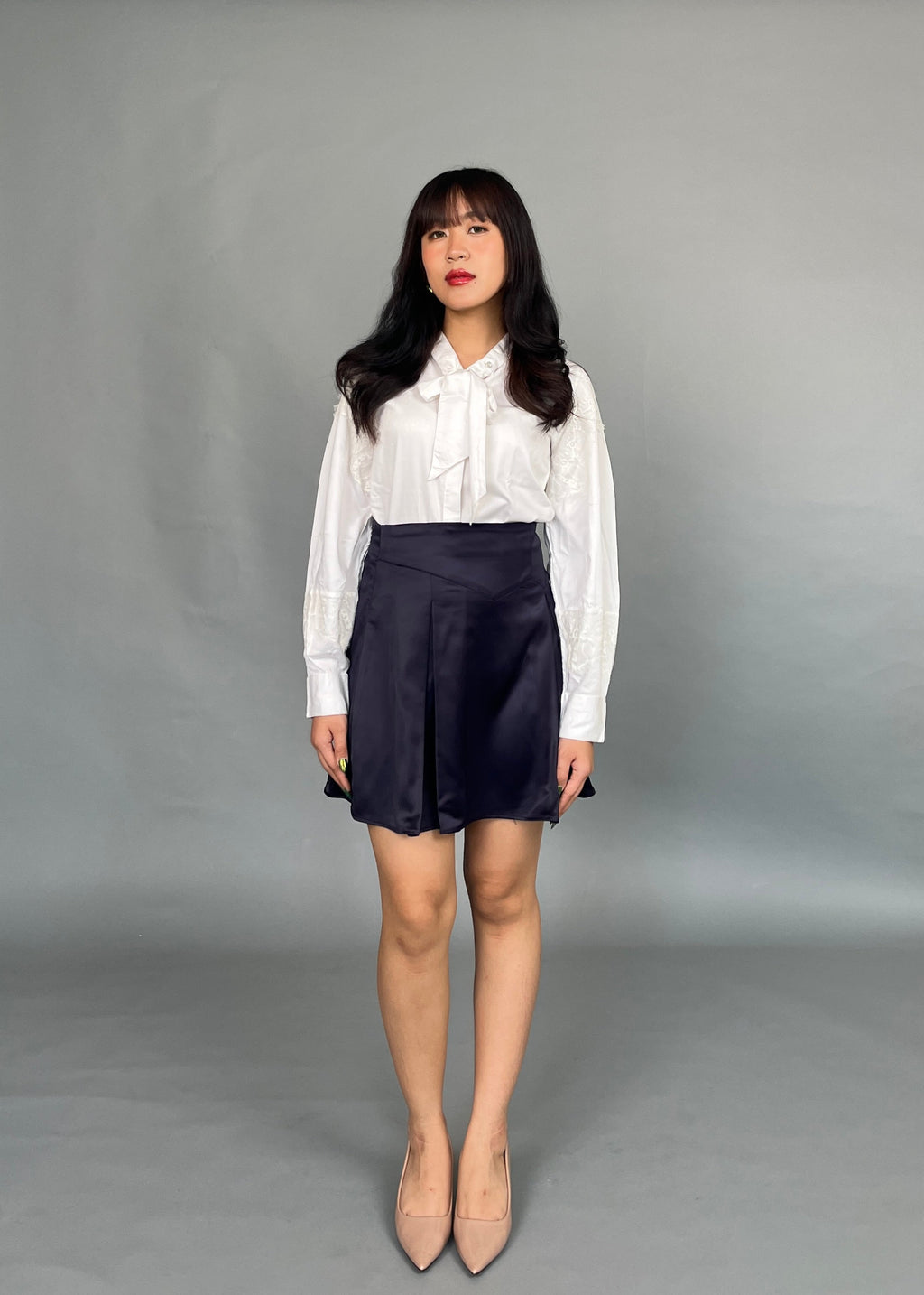 A woman wearing a white Dorothea lace shirt with lantern sleeves and a neck-tie, paired with a dark skirt and beige shoes.