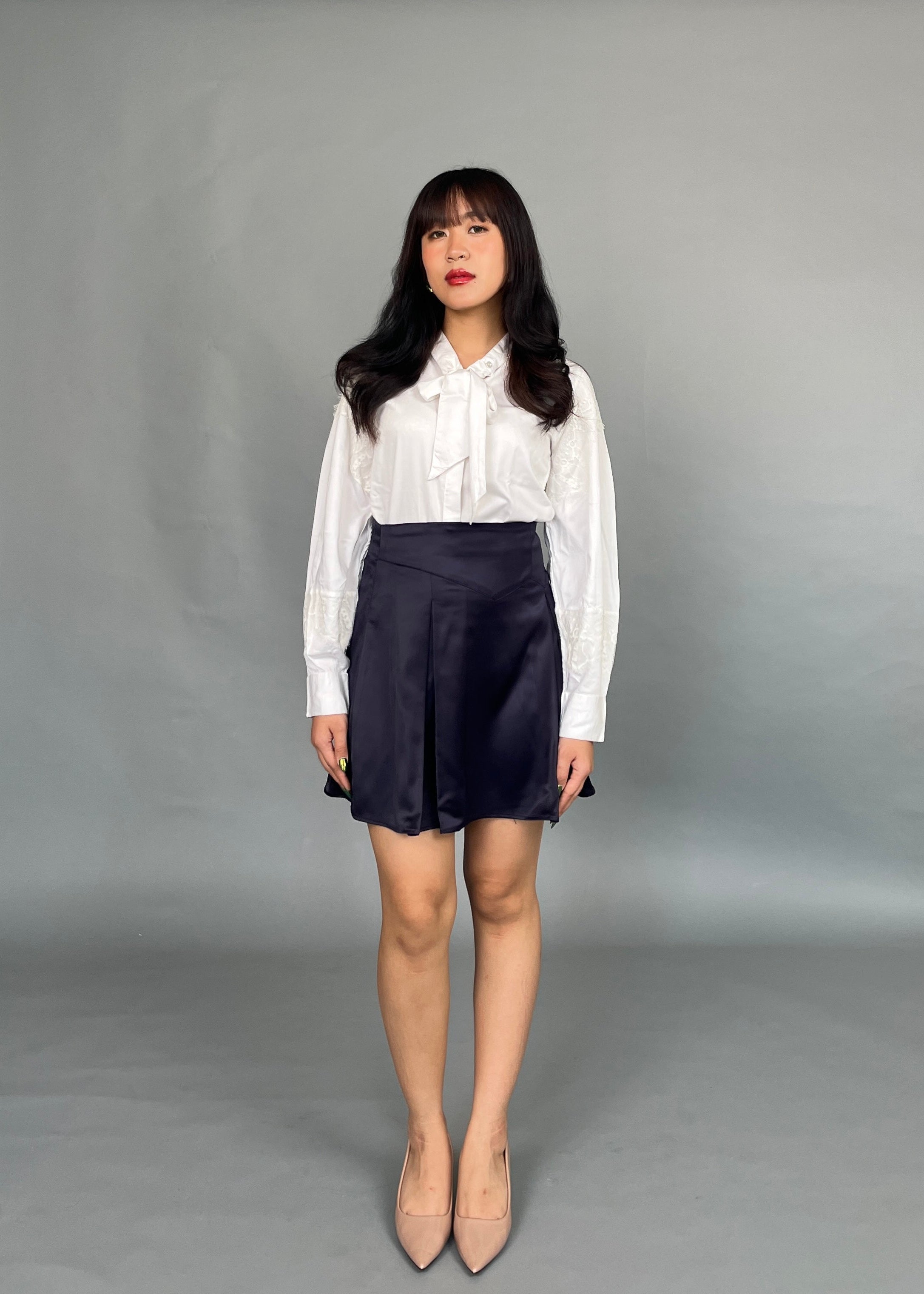 A woman wearing a white Dorothea lace shirt with lantern sleeves and a neck-tie, paired with a dark skirt and beige shoes.