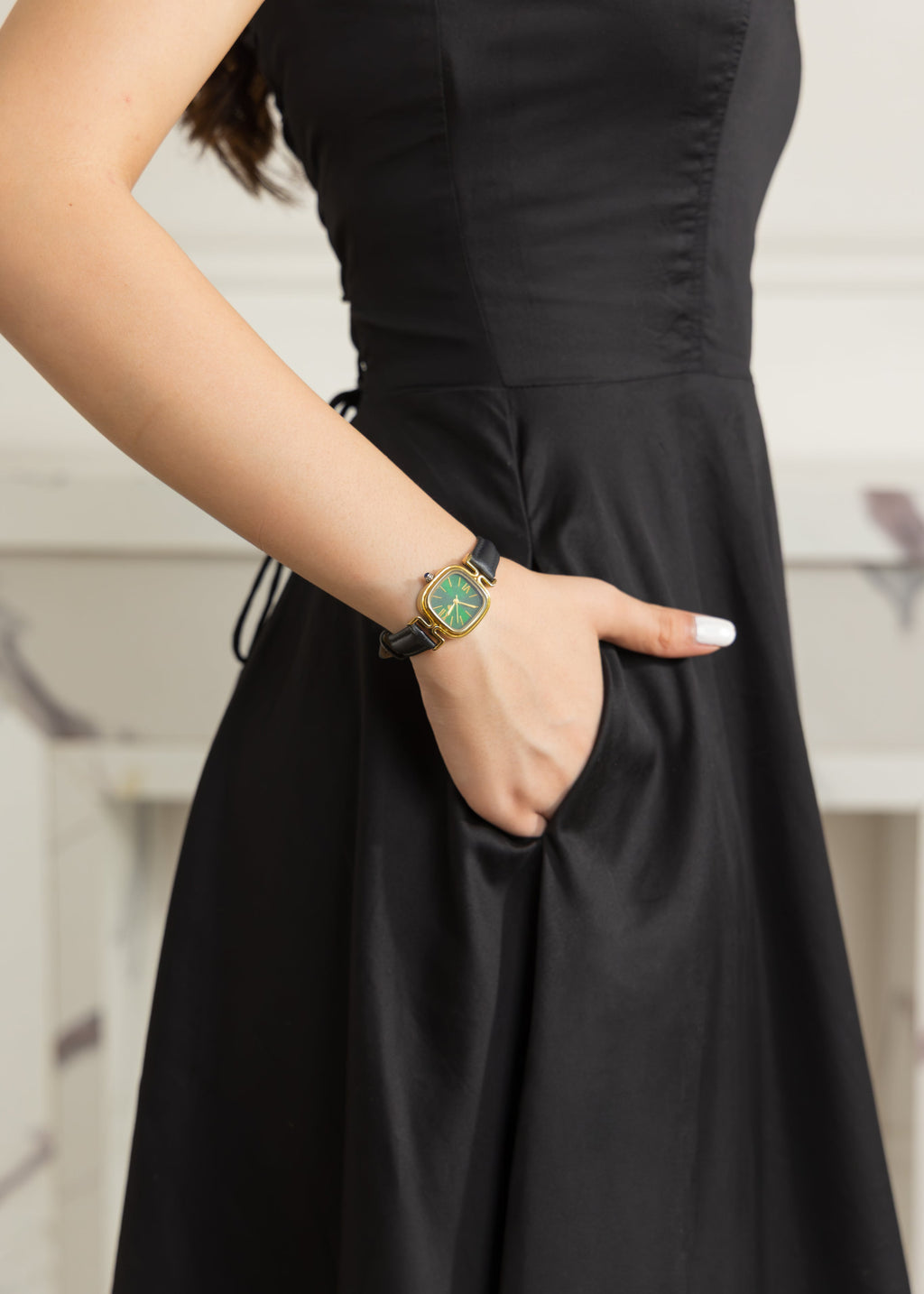 A close-up of a woman wearing a black sleeveless dress and a gold watch with a green dial, with her hand in her pocket.