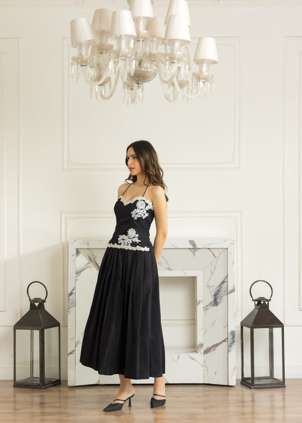 A woman models a black midi dress with white floral embroidery on the bodice and waist, standing in a bright room with a marble fireplace, chandelier, and lanterns.