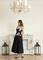 A woman models a black midi dress with white floral embroidery on the bodice and waist, standing in a bright room with a marble fireplace, chandelier, and lanterns.