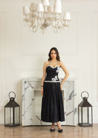A woman stands in a grand room with a marble fireplace, chandelier, and lanterns, wearing a black strapless midi dress with white floral embroidery on the bodice and waist.