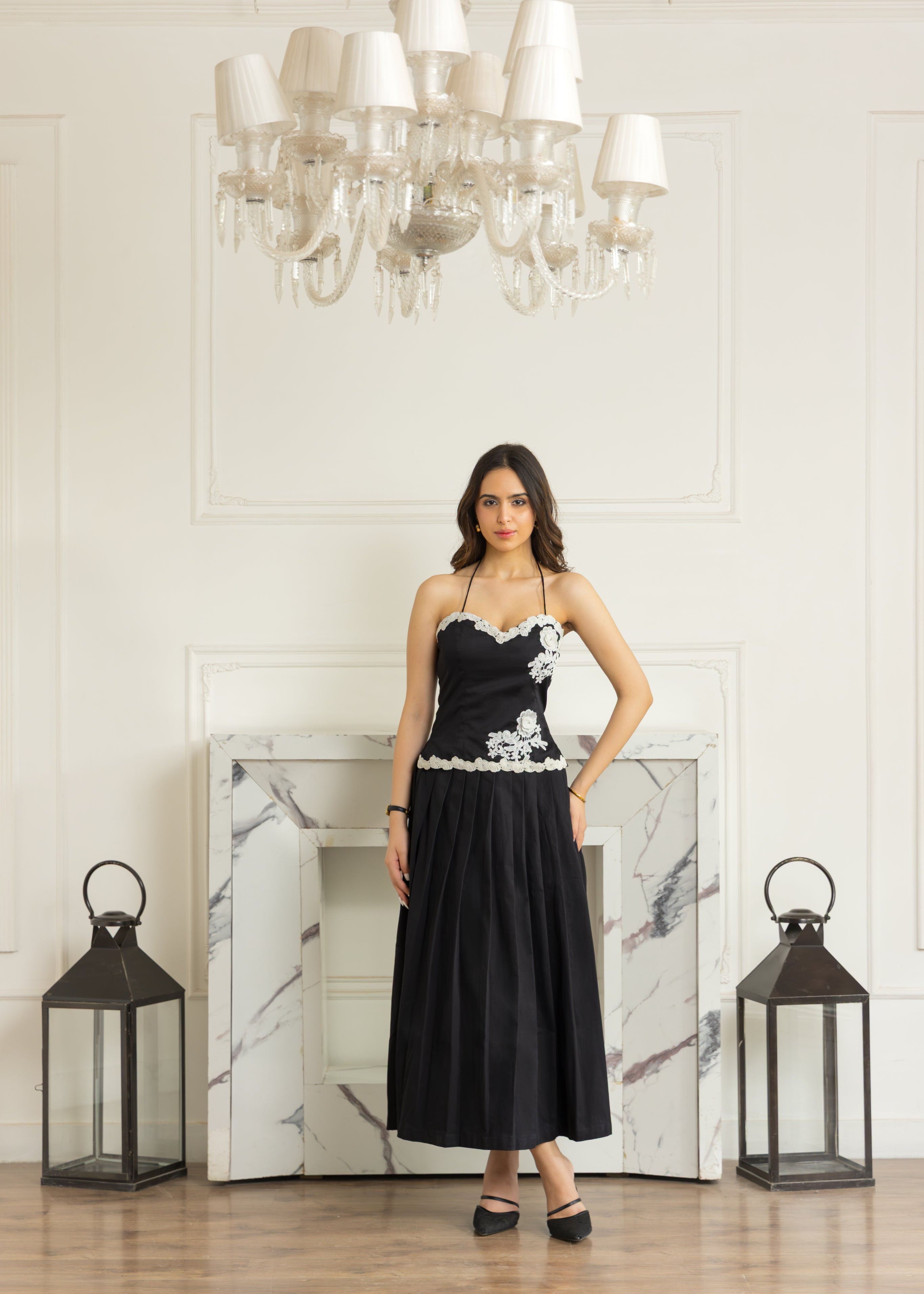 A woman stands in a grand room with a marble fireplace, chandelier, and lanterns, wearing a black strapless midi dress with white floral embroidery on the bodice and waist.