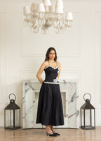 A woman stands in a bright room wearing a black midi dress with white lace trim at the bust and waist. The room features a white paneled wall, a marble fireplace, a large chandelier, and two black lanterns on the floor.
