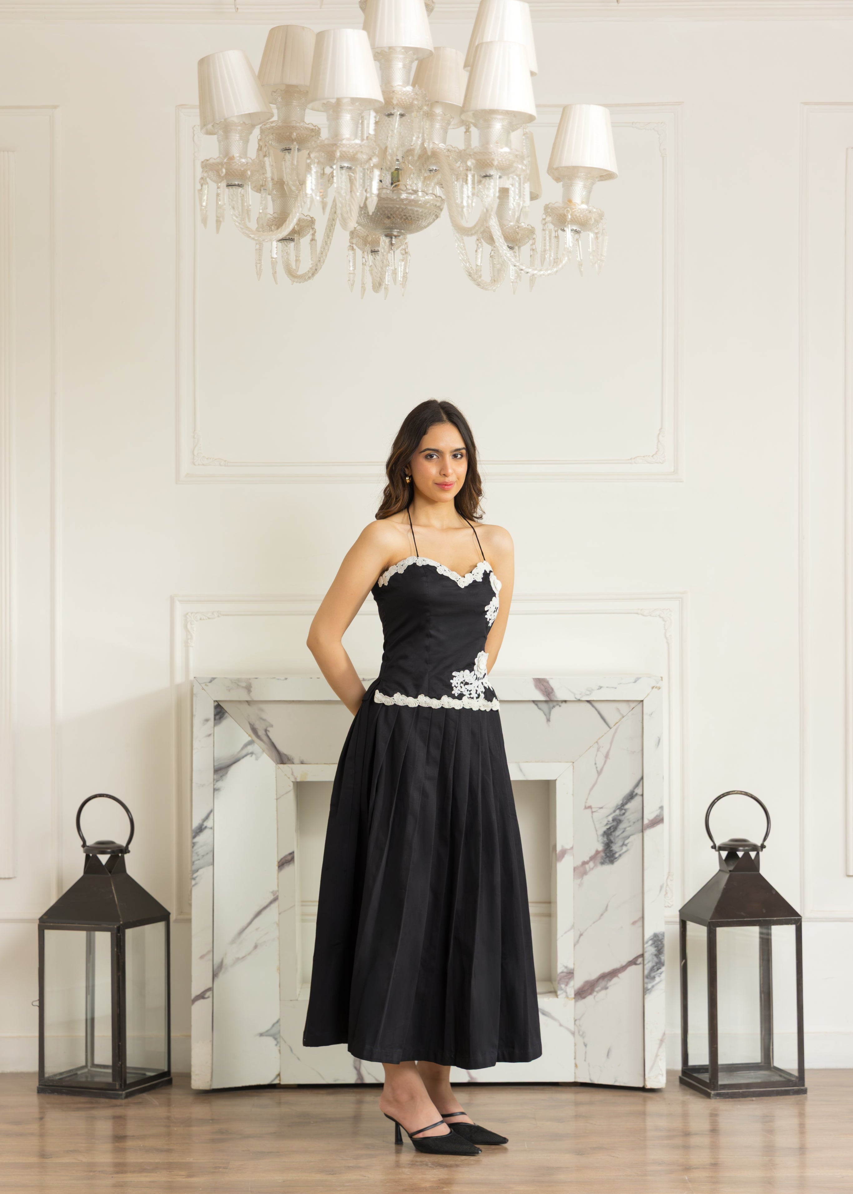 A woman stands in a bright room wearing a black midi dress with white lace trim at the bust and waist. The room features a white paneled wall, a marble fireplace, a large chandelier, and two black lanterns on the floor.