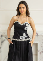 A woman modeling a black halter-neck dress with white floral lace appliques and a pleated skirt, standing in front of a white marble fireplace.