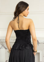 A woman shown from the back wearing a black strapless smocked dress with tie-back neckstrings and a ruffled waist, photographed indoors against a light wall and marble fireplace.
