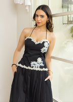 A woman wearing a black halter-neck dress with white floral lace appliques and a pleated skirt, standing in an indoor setting.